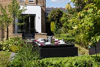 Black dining table and chairs on patio surrounded by flowerbeds in garden.