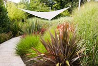Contemporary garden incorporates curving stone path through beds planted
 with Imperata cylindrica 'Rubra', Phormium 'Sundowner', 
Pennisetum alopecuroides 'Cassians' Choice' to area with suspended awning
 