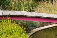 Wooden bridge across pond near ornamental grasses such as Festuca glauca 'Elijah Blue' and Chasmanthium latifolium
