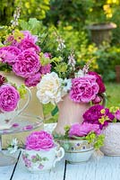 Cut roses arranged in enamel tea set, against a garden backdrop.