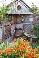 View over flowering Crocosmia to sheltered seating area with wooden chair and table in cottage garden. Hilltop, Stour Provost, Dorset, UK.