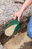 Woman using plastic scoop to add gravel and sand in hole dug for firepit. 