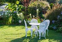 White meal table and chairs on the lawn surrounded by flowering borders. 