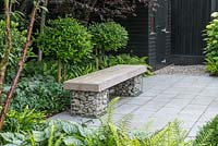 A wooden bench supported on gabions of pebbles, on a shady granite patio.