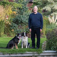 Man with two dogs on lawn in a garden full of plants
