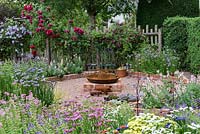 Enclosed garden with brick-edged beds of flowers, Antirrhinum majus - 
 snapdragon, Limonium - statice and Slavia - clary sage, a wooden fence 
covered in Rosa - roses - and Clematis. In centre is a focal point based around iron pig feeding trough.