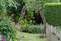 An ornate arch straddles a grass path, framing a view of two hares crafted 
from chickenwire