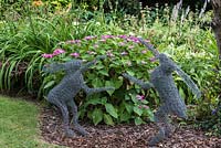 Two dancing hares, crafted from chickenwire, in front of flower border