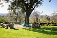 A holm oak, Quercus ilex, is encircled by a metal bench and balustraded terrace on the lawn in front of the house at Plas Brondanw, Penrhyndeudraeth, Gwynedd, Wales