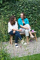 View of two women chatting on wooden bench in garden. 