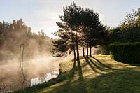 Mist rising from the lake with metal sculpture at sunrise. Plaz Metaxu Garden, Devon, UK.