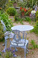 Ornate table and chairs in gravelled terrace, surrounded by flowering perennials and potted plants. 