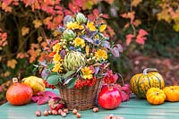 Autumnal floral arrangement in wicker basket, surrounded by harvested vegetables, fruit and nuts.