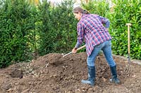 Woman adding soil over pile of wood, garden waste and leaf matter, to create a raised mound in empty bed.
