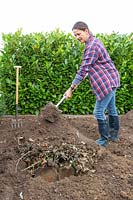 Woman adding soil over pile of wood, garden waste and leaf matter, to create a raised mound in empty bed