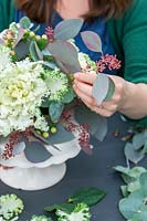 Woman adding seeded Eucalyptus to arrangement with ornamental cabbage.