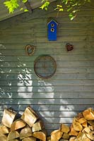 Log store to the rear of the summer house with bird house, woven willow hearts and old garden sieve. 