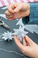 Woman using needle and silver thread to create hanging loop on paper star decoration.