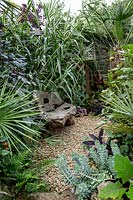 Step down from the lawn leads to gravel area and seat surrounded by exotic foliage planting. 
