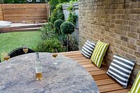 View across modern outdoor table and wooden seating dressed with garden cushions against brick wall
 to an artificial lawn with Ligustrum jonandrum trained as half standards to horizontal wooden fencing