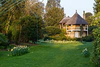 View over lawn with drifts of daffodils to Swiss-style thatched cottage with trees nearby
 