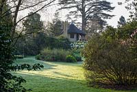 View over lawn to island beds with mix of shrubs and perennials towards thatched cottage