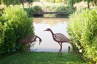 Metal bird sculptures next to lake and wooden bridge beyond. Bowley Farm, Sussex, UK. 