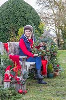 Woman sat on bench holding homemade wreath, surrounded by Christmas greenery and decorations