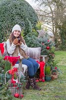 Woman sat on bench holding warm drink, surrounded by Christmas greenery and decorations