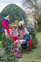 Woman giving basket with presents to friend sat on bench, surrounded by Christmas greenery and decorations