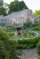 Nymphaea waterlilies in a circular pond with stone fountain surrounded by Buxus - box hedging. Rare Lean-to greenhouse.  Rosa 'Queen of Sweden' and Nepeta grandiflora 'Summer Magic' in front of greenhouse