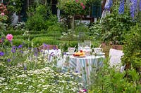 Table set for tea and cake in garden surrounded by flowering shrubs and perennials. 
