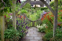 View from under wooden pergola to low gate and flowering herbaceous perennial borders. Wollerton Old Hall, Market Drayton, UK. 
