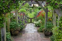 View from under wooden pergola to low gate and flowering herbaceous perennial borders. Wollerton Old Hall, Market Drayton, UK.