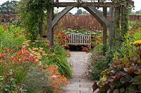 Double, mixed flowerbeds bordering paved pathway, with view to traditional wooden bench. Wollerton Old Hall Garden, Market Drayton, UK.