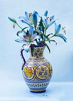 Dyed blue oriental cut lilies in decorative vase, set against blue background.