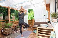 View of mediterranean summer balcony. White curtains as Sun protection and blinds.Yoga place with mat and Buddha, decorated with rose petals. Women in Vrksasana position.