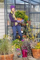Woman carrying potted evergreen shrub out of greenhouse.