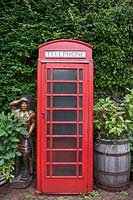 Traditional red telephone box and wooden Native American chief sculpture on brick-paved driveway, surrounded by oak barrels of Amaranthus caudatus - Love Lies Bleeding.
