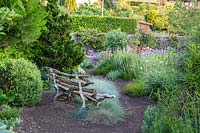 View from a rustic wooden bench with ornamental grass Festuca glauca nearby towards bed 
of flowering perennials. Behind bench evergreens such as 
Chamaecyparis lawsoniana 'Erecta Aurea'.