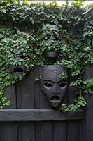 A black painted timber fence decorated with three Aztec style black wooden masks that have Ficus pumila, - Creeping Fig - growing over them.