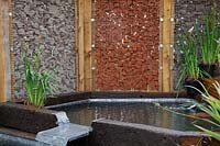 Pond with metal waterfall with gabion-style walling as a windbreak in 
RHS Garden for a Changing Climate show garden 