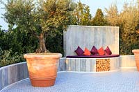The Retreat, show garden featuring mature Olive trees - Olea europaea -
 in terracotta pots,  and a backdrop of Bay - Laurus nobilis. 
Ceramic tiling on the floor and a lime-washed wooden semi-circular seat
 with purple cushions which doubles as a decorative log store for the heated spa.