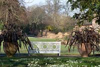 A white garden bench flanked by terracotta pots planted with Phormium tenax 'Atropurpureum' - Bronze New Zealand Flax.