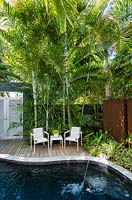 Set of garden chairs and table on wooden decking by pool, surrounded by tropical palms. Von Phister Residence, Key West, Florida, USA. Garden design by Craig Reynolds.
