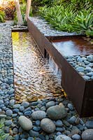 View along a water feature of corten steel filled with cobbles, a waterfall and rill with Agave weberi plants at back
