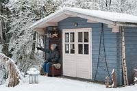 A woman sitting in front of blue summerhouse covered by snow in winter.
