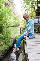 Woman sitting on a deck with her bare feet dangling in the water of a creek
