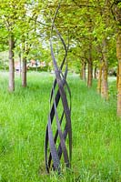 Flame - sculpture in steel - set in grass with avenue of trees beyond
