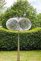 Torodial - sculpture in stainless steel -  on lawn with hedge and trees beyond
